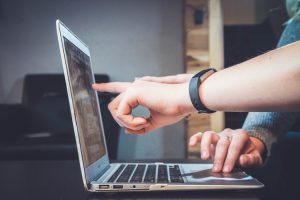 A person's hand touching the touchpad from a laptop and anothe person's hand pointing to the screen